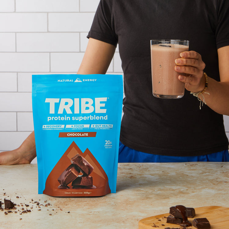 Person holding a glass of chocolate protein shake next to a bag of Tribe Protein Superblend on a wooden surface.