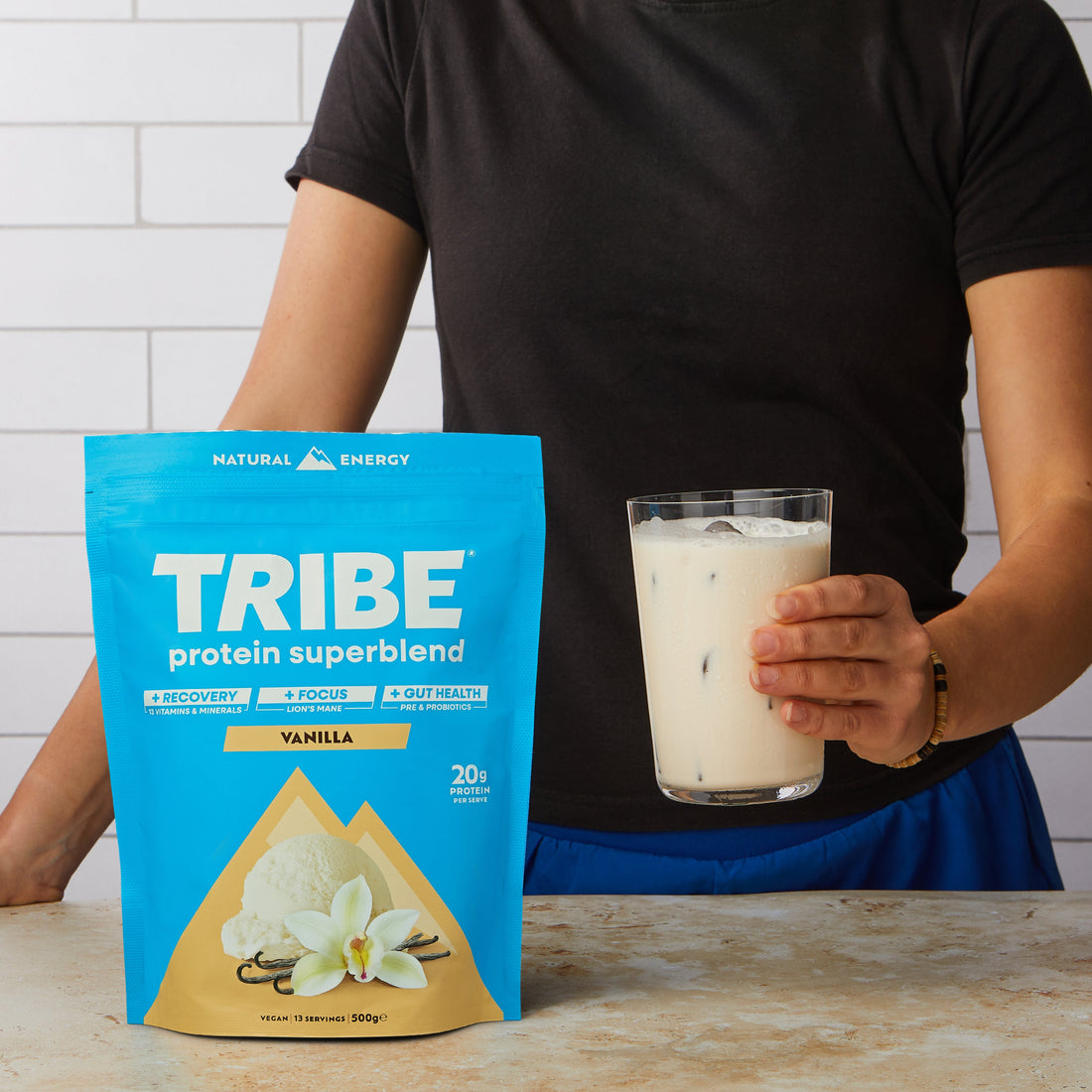 Women holding a glass of vanilla Protein Superblend by Tribe, with a pouch on the table in front of her.