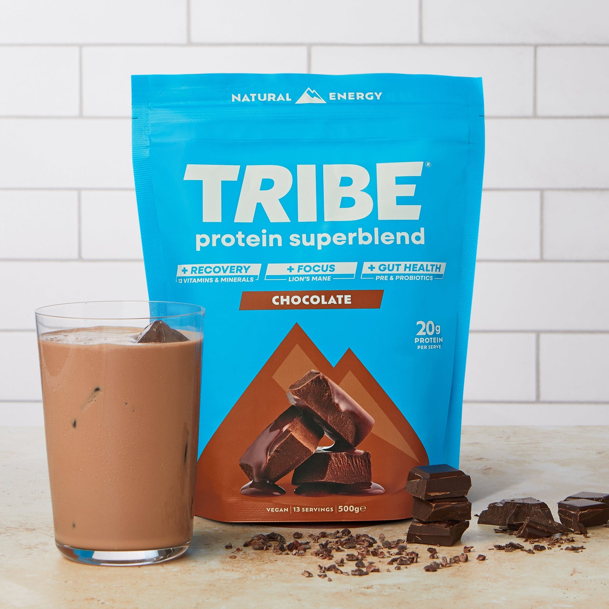 TRIBE protein superblend chocolate flavour pouch with a glass of chocolate shake on a wooden surface, with chocolate pieces on the surface.
