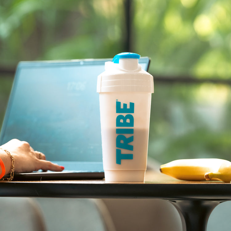 Person using a laptop with a 'TRIBE' shaker bottle and banana on a table.